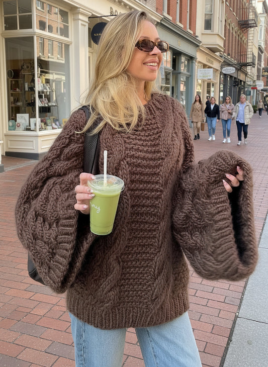 Woman wearing a brown chunky knit sweater holding a green drink outdoors.