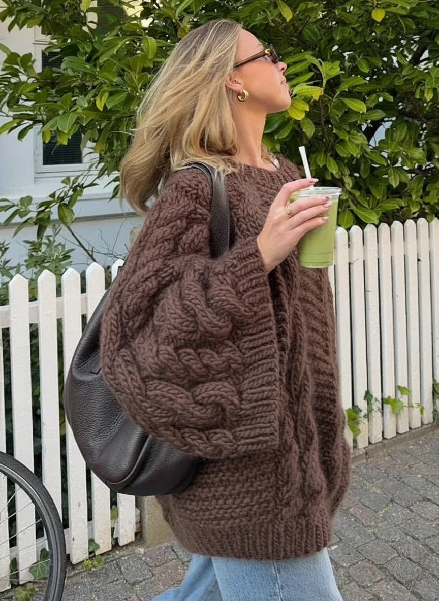 Woman in a brown cable knit sweater drinking from a green iced drink outdoors.